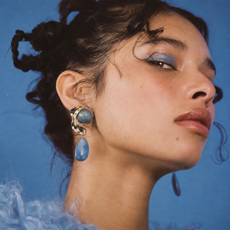 blue drop earrings beauty close-up product photo: A close-up editorial portrait shows a model wearing sculptural gold-and-blue drop earrings against a vivid bl…