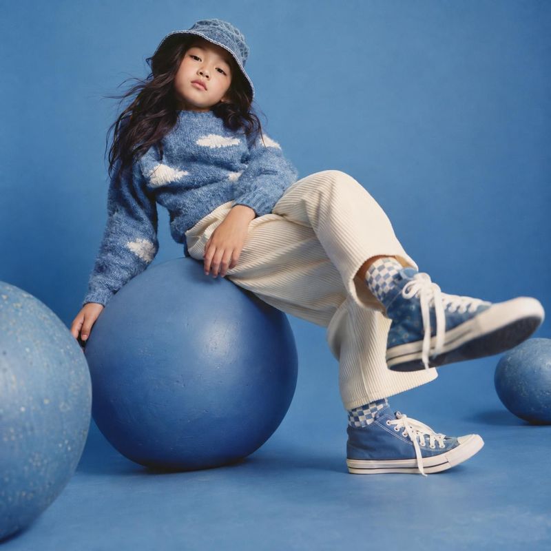 children's clothing and sneakers model try-on product photo: A child in a blue cloud-pattern sweater, cream trousers, matching sneakers, and a bucket hat poses…