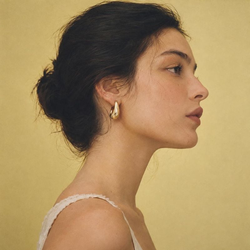 gold hoop earrings model try-on product photo: A woman in profile wears sculptural gold earrings and a delicate white top against a warm yellow studio backdrop…