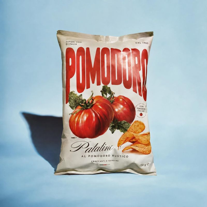 tomato-flavored potato chips food and drink product photo: A cream-colored bag of tomato-flavored potato chips stands upright on a pale blue studio surface, wi…