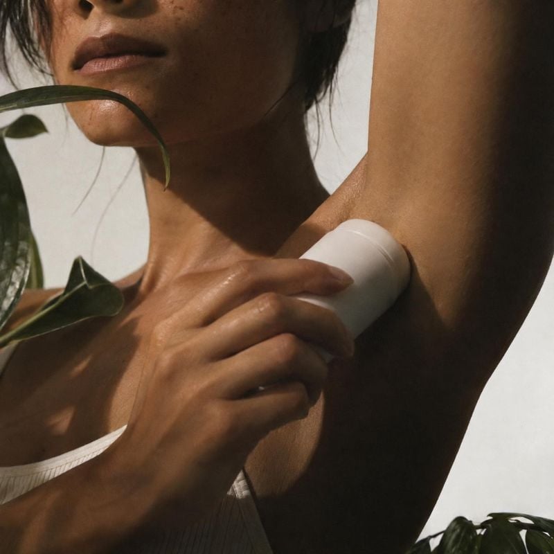 deodorant stick model try-on product photo: A tightly cropped woman applies a white deodorant stick beneath her arm, with leafy greenery and a pale backdrop ad…