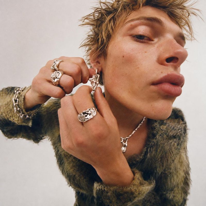 silver jewelry model try-on product photo: A close, slightly wide-angle studio portrait shows a young man adjusting a silver earring, styled with chunky rings,…