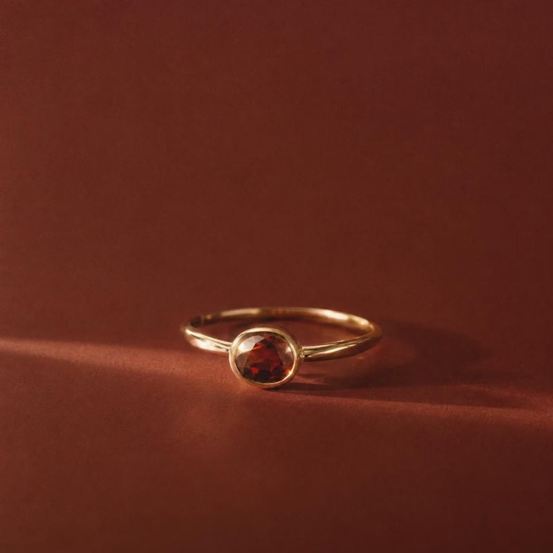 gold ring with a red gemstone close-up detail product photo: A delicate gold ring set with a deep red gemstone rests on a softly textured rust-colored surface,…