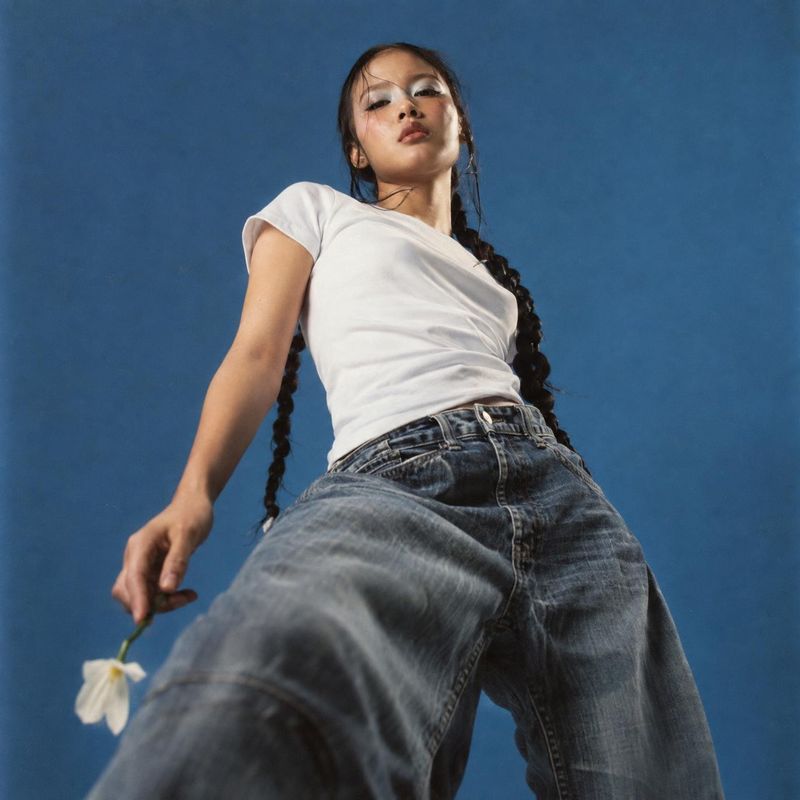 jeans model try-on product photo: A model in a white T-shirt and loose blue jeans poses from a dramatic low angle against a blue studio backdrop, holding a sma…