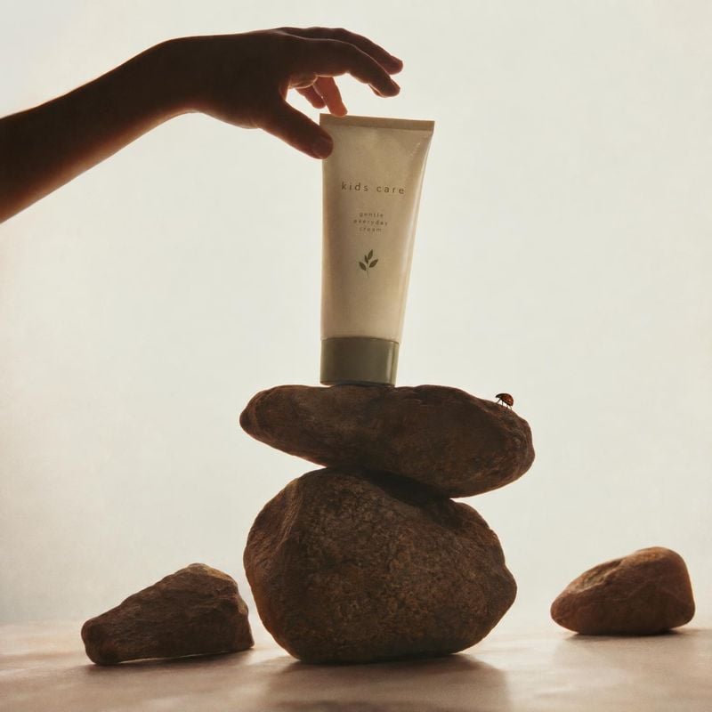 kids' everyday cream product in hand product photo: A hand reaches in to present a minimalist kids' cream tube atop stacked stones against a softly lit neutral…