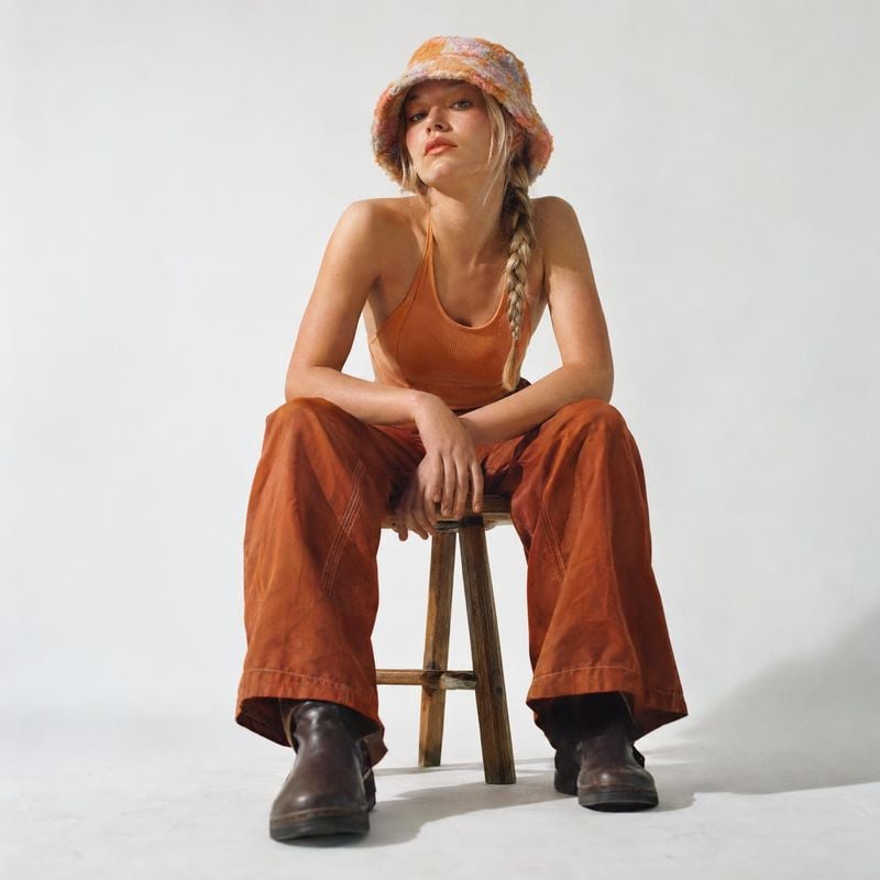 women's outfit model try-on product photo: A woman in a rust-colored outfit, dark boots, and a patterned bucket hat sits on a wooden stool against a softly lit…