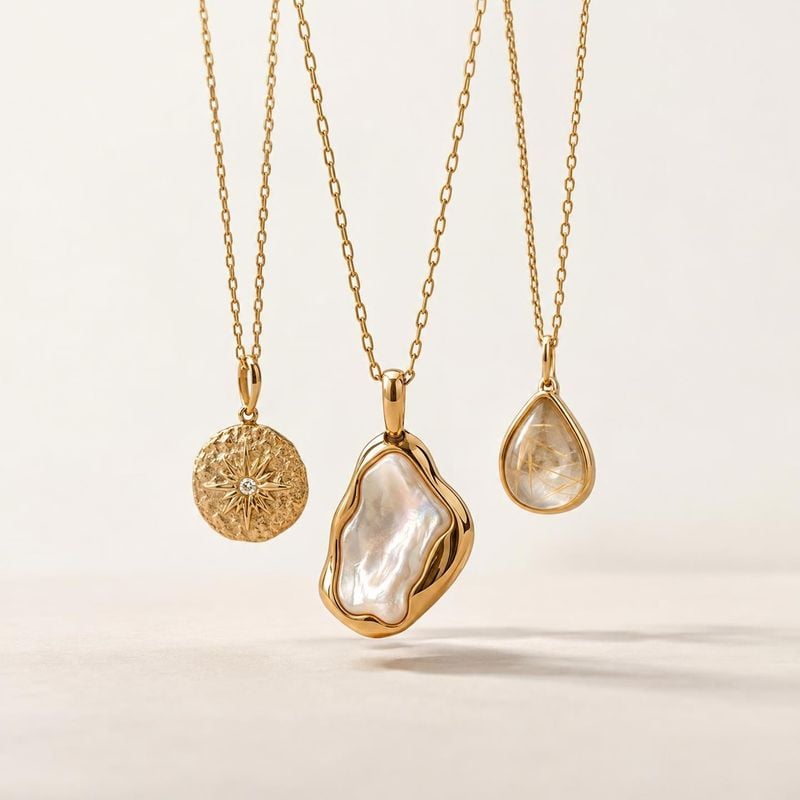 gold pendant necklaces group lineup product photo: Three gold-tone pendant necklaces hang against a warm cream studio backdrop, their polished metal and irides…