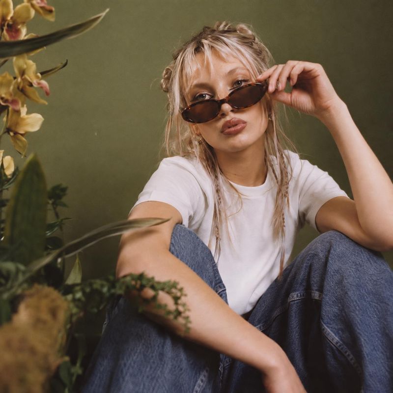sunglasses model try-on product photo: A woman in a white T-shirt and blue jeans poses in dark sunglasses against an olive-green backdrop with foliage, in soft…