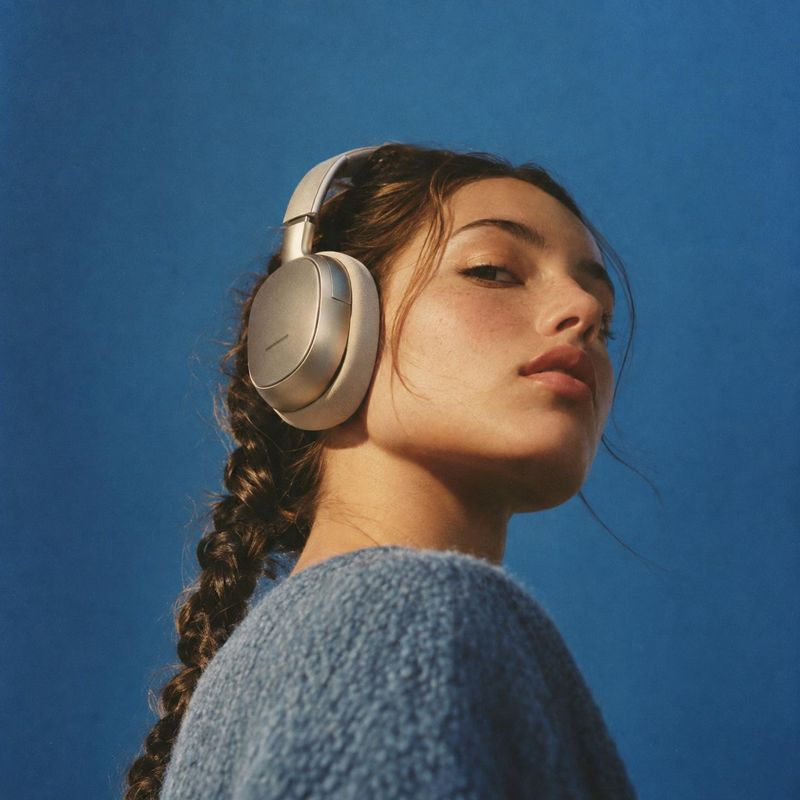 over-ear headphones model try-on product photo: A woman in a textured blue sweater wears sleek silver over-ear headphones against a vivid blue studio backdrop,…
