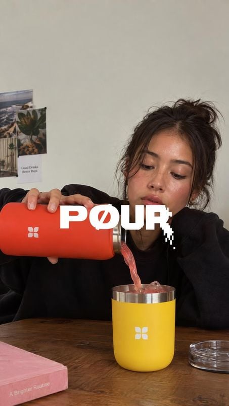 insulated tumbler splash / pour product photo: A woman in a black sweatshirt pours a pink drink from a red tumbler into a yellow one at a wooden table, with bo…