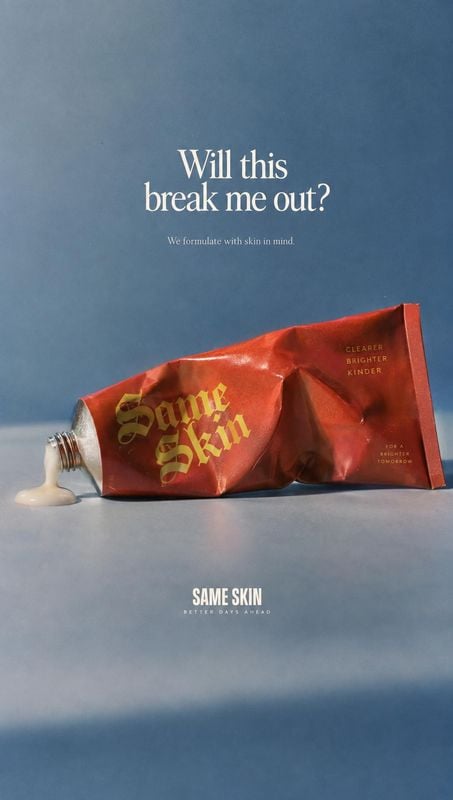 skincare tube still life product photo: A crumpled red skincare tube spills a small dollop of cream across a blue studio backdrop, with centered white and gold…