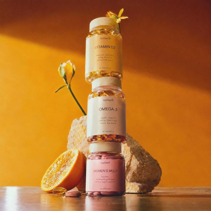 vitamin supplements personal care product photo: Three labeled supplement bottles are stacked among a citrus half, a flower, and pale stone against a warm oran…