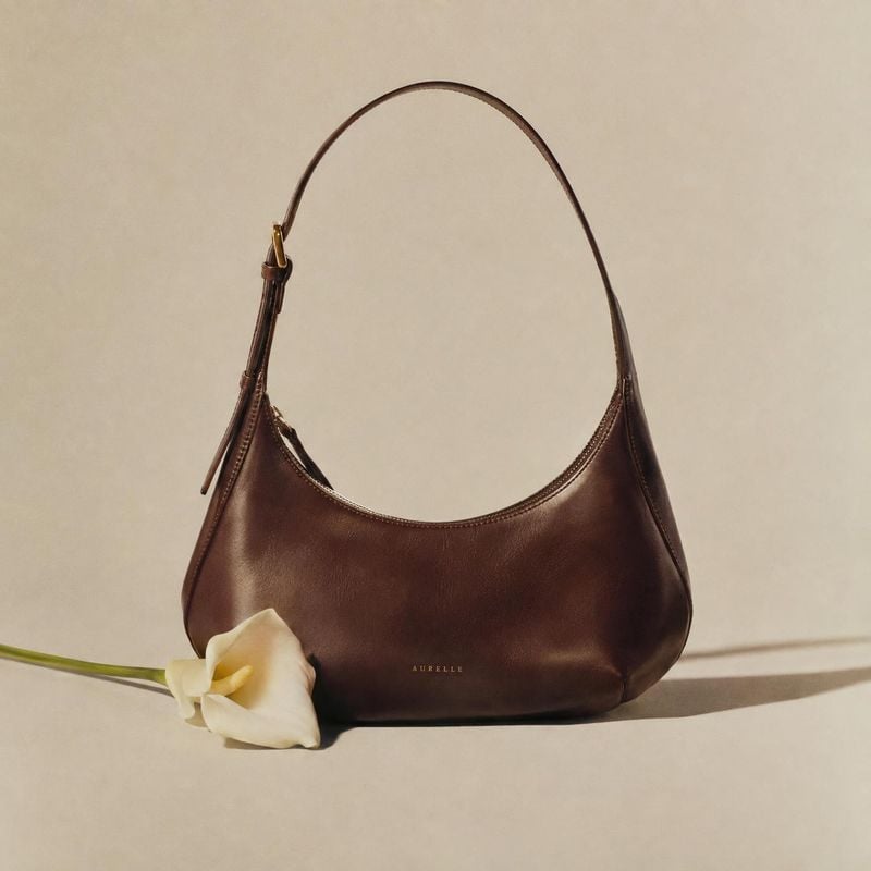 leather shoulder bag fashion product photo: A dark brown leather shoulder bag rests on a warm beige studio surface beside a white calla lily, lit softly to cre…