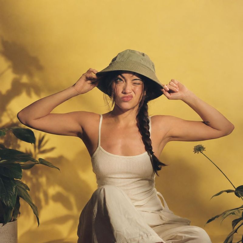 bucket hat model try-on product photo: A woman in a neutral outfit and olive bucket hat poses against a warm yellow studio backdrop with leafy plants, lit by s…