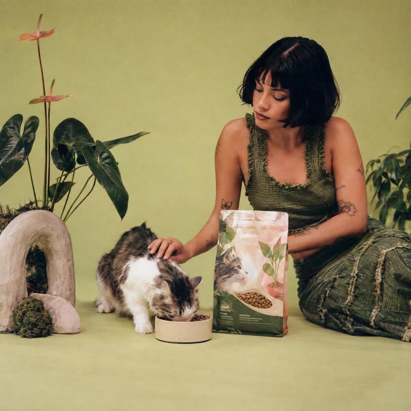 cat food lifestyle scene product photo: A woman in a textured green outfit gently pets a cat eating beside its food bag, styled with leafy plants against a sof…