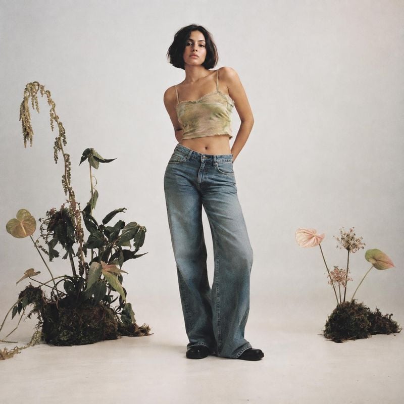 wide-leg jeans model try-on product photo: A model poses in wide-leg jeans and a muted crop top against a softly lit studio backdrop, framed by potted plants f…