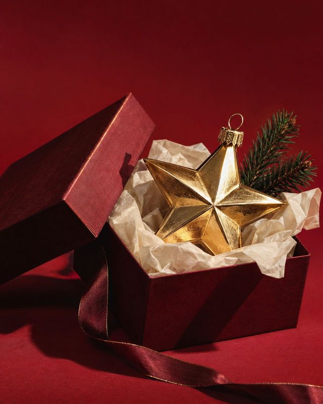 gold star Christmas ornament christmas product photo: A faceted gold star ornament rests in tissue paper inside an open burgundy gift box, with evergreen sprig…