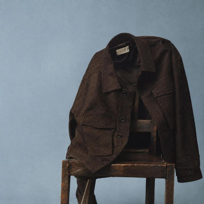 dark brown jacket fashion product photo: A dark brown textured jacket is draped over a wooden chair against a cool blue-gray studio backdrop, with soft directi…