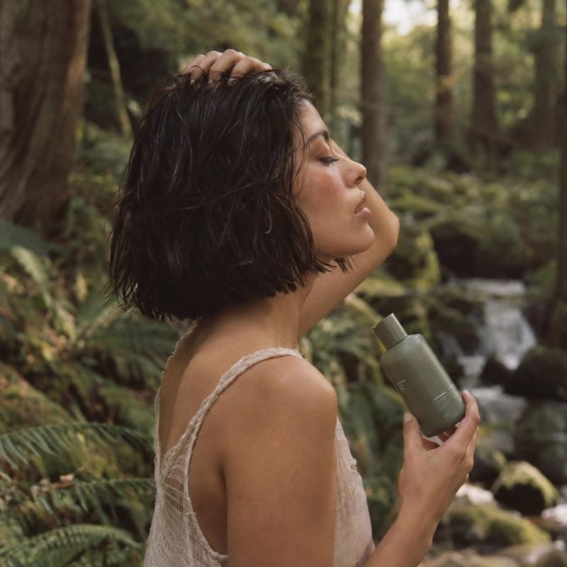 hair-care bottle lifestyle scene product photo: A woman with short, wet hair holds a muted green hair-care bottle beside a forest stream, with soft natural lig…