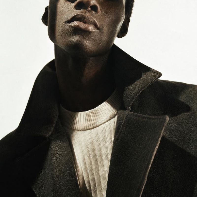 wool coat close-up detail product photo: A tightly framed male fashion model wears a dark wool coat over a cream ribbed sweater against a pale studio backgroun…