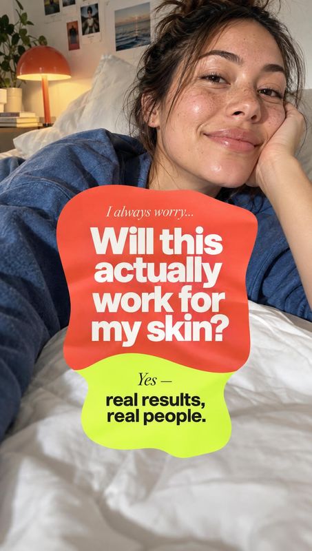 beauty close-up product photo: A woman reclines on a bed in a casual blue top, smiling softly in warm bedroom light beneath bold red-and-lime skincare messagin…