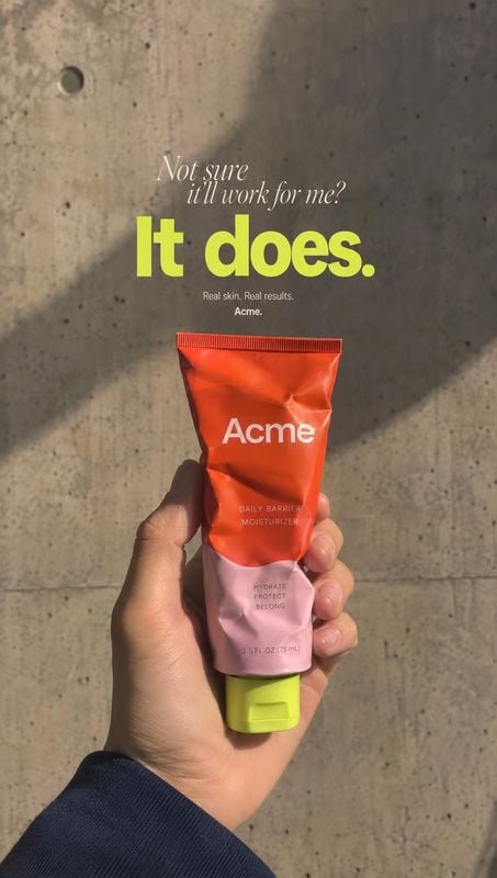 moisturizer tube product in hand product photo: A hand presents a bright red-and-pink moisturizer tube against a sunlit concrete wall, with bold lime-green pro…