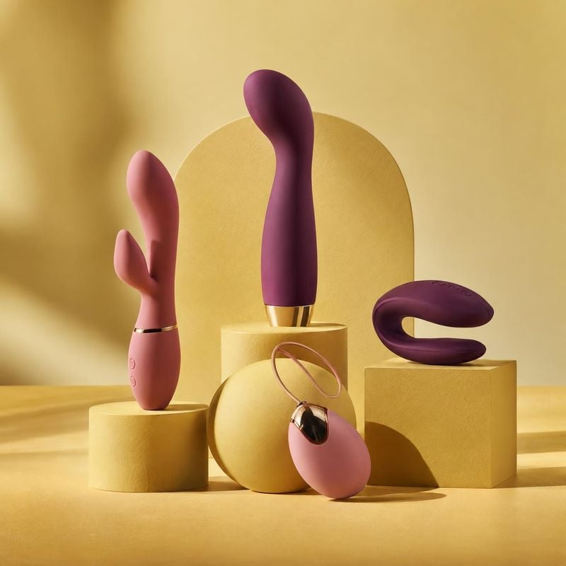 sexual wellness devices group lineup product photo: A coordinated set of pink and plum personal wellness devices sits on sculptural yellow pedestals in warm, d…