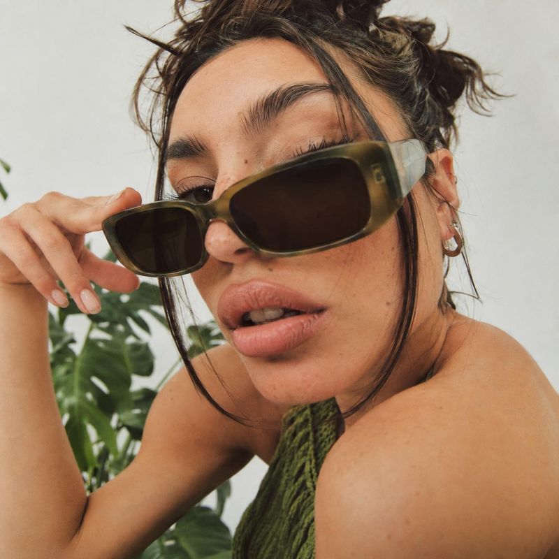 sunglasses model try-on product photo: A close-cropped fashion portrait shows a woman wearing chunky olive-toned sunglasses and a textured green top, softly li…
