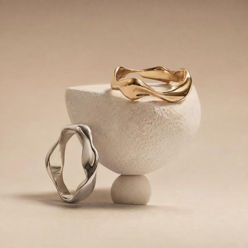 sculptural rings jewelry product photo: Two sculptural gold- and silver-toned rings rest on pale stone forms against a warm neutral studio backdrop, lit softly…