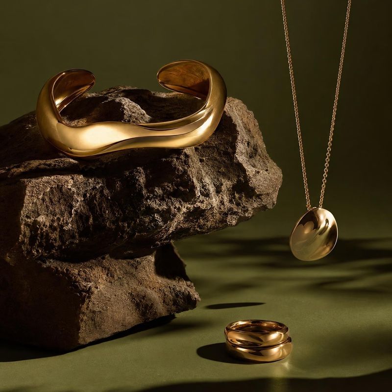 gold jewelry set jewelry product photo: A sculptural gold bracelet, pendant necklace, and ring are arranged among rough rocks against an olive-green studio bac…