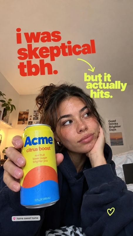 canned citrus drink product in hand product photo: A young woman in a dark hoodie holds a bright yellow citrus drink can toward the camera in a cozy home inter…