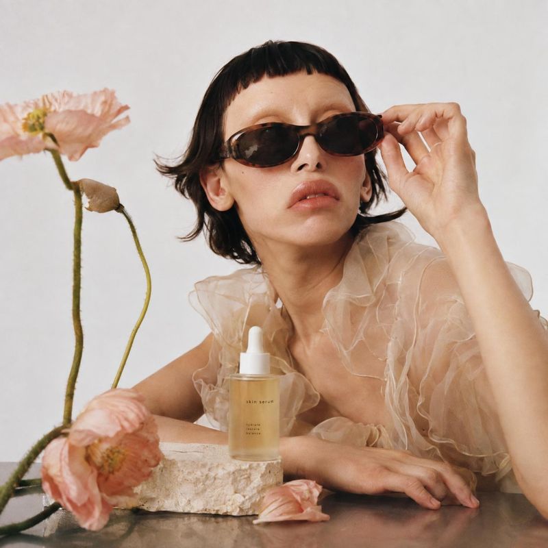 skin serum beauty close-up product photo: A short-haired model in dark sunglasses and a sheer ruffled top poses beside a serum bottle and soft pink flowers aga…