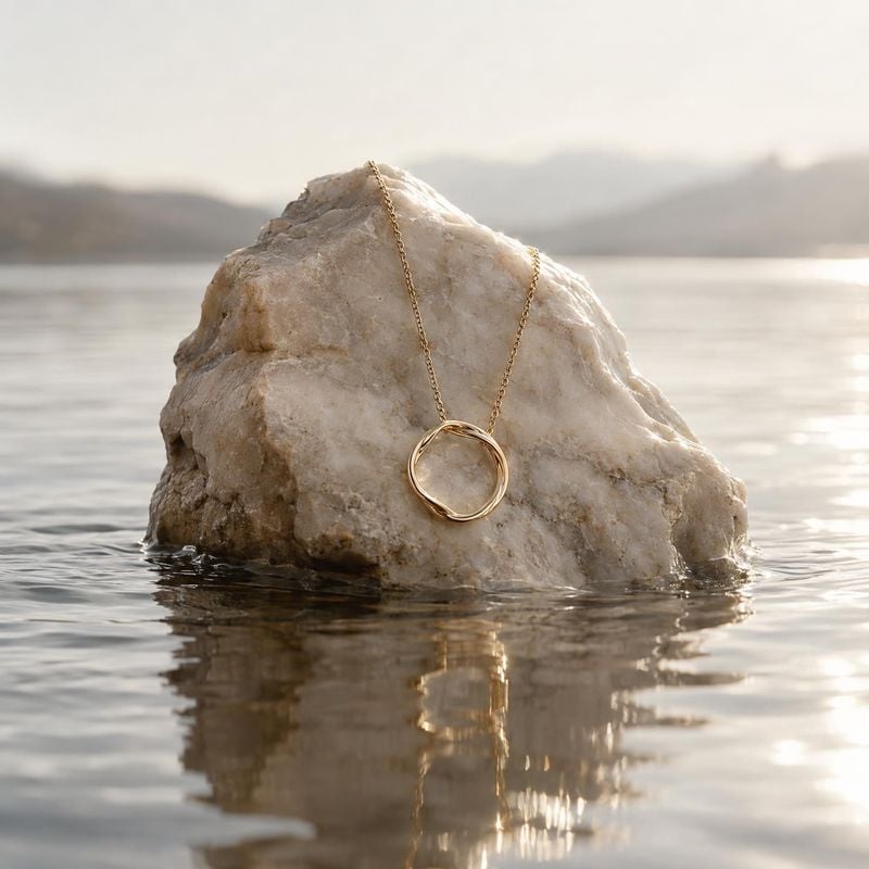gold pendant necklace jewelry product photo: A delicate gold pendant necklace drapes over a pale rock rising from calm water, lit by soft golden-hour light for…