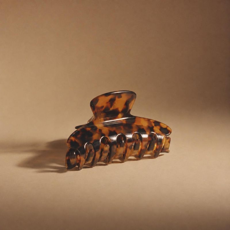 tortoiseshell hair claw clip jewelry product photo: A glossy tortoiseshell-pattern hair claw clip rests on a warm beige surface, softly lit with a long shadow…