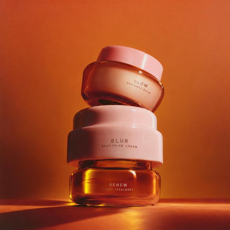 skincare cream jars group lineup product photo: Three pink and amber skincare jars are stacked against a warm orange studio backdrop, with glossy highlights an…