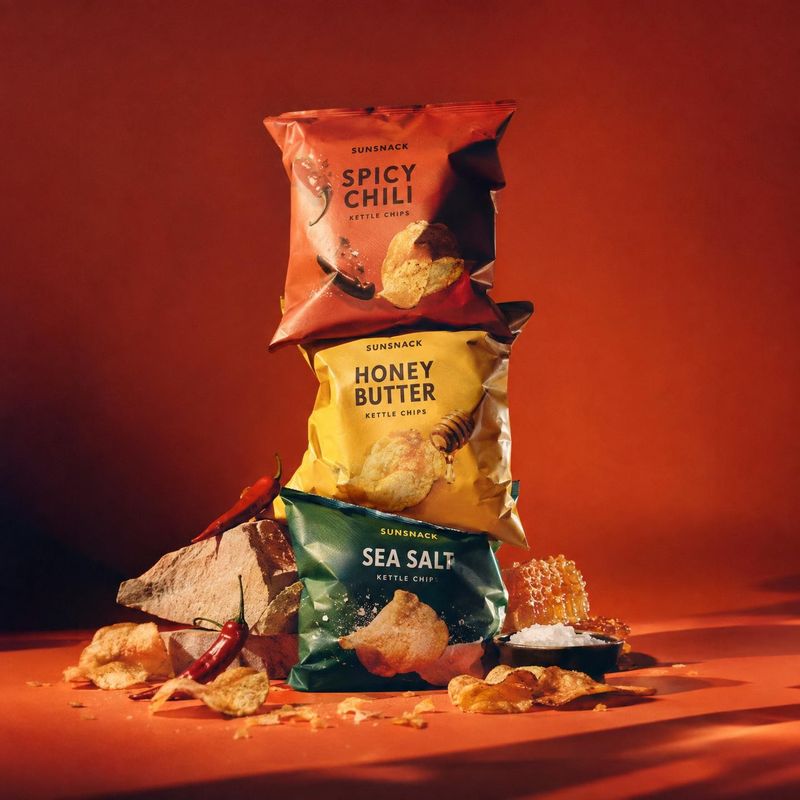 kettle chips group lineup product photo: Three colorful kettle-chip bags are stacked among chips, chilies, honeycomb, and salt on a dramatic red studio set, li…