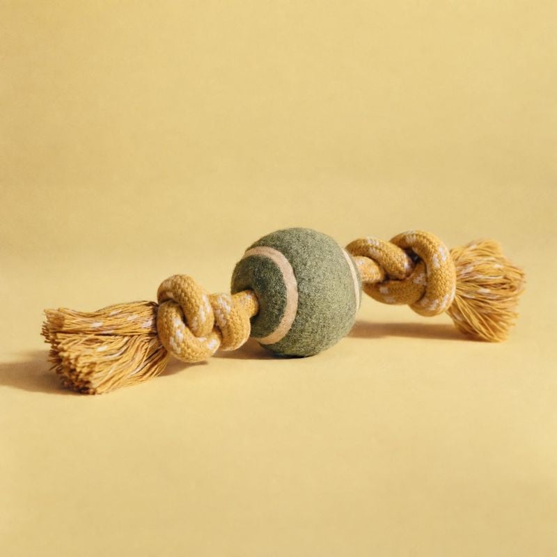 rope-and-ball dog toy pet product photo: A braided rope dog toy with tasseled ends and a felt ball sits centered on a warm beige studio backdrop, lit softly fo…