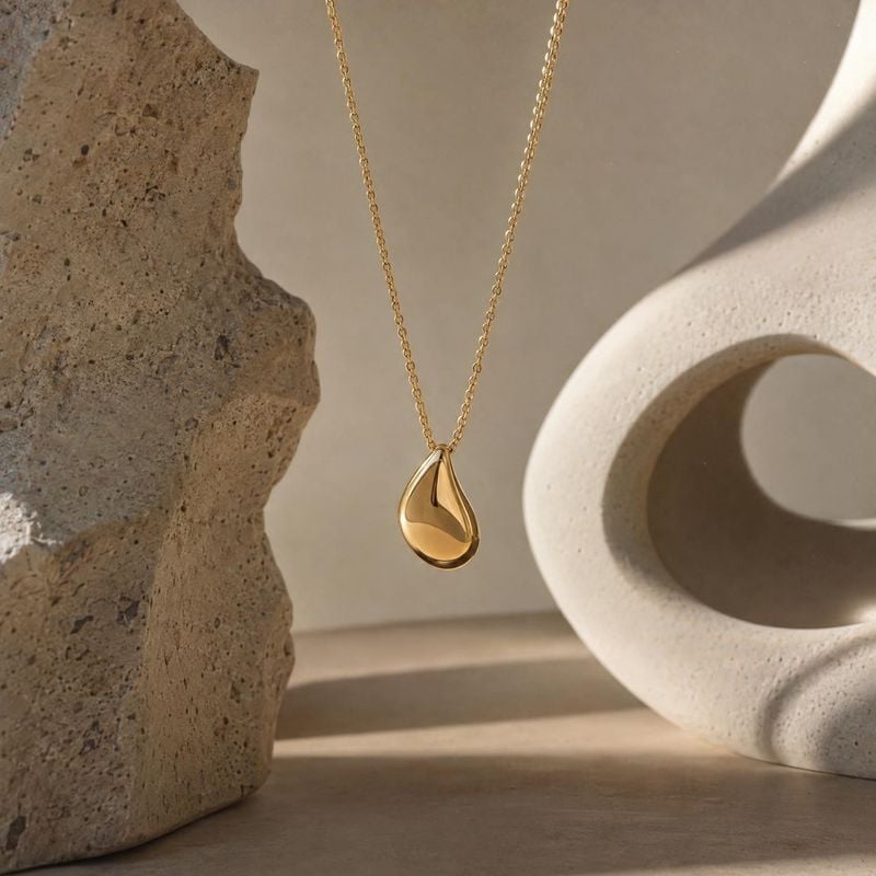 gold pendant necklace jewelry product photo: A polished gold teardrop pendant hangs between sculptural stone forms in warm, directional light, creating a refin…