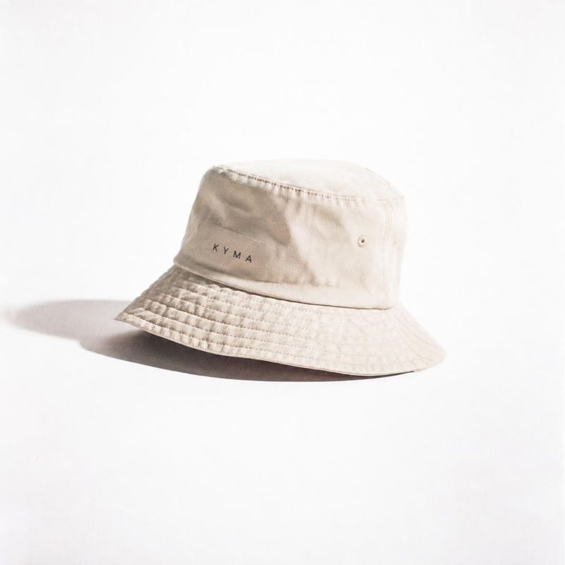 beige bucket hat fashion product photo: A beige bucket hat with small dark lettering sits alone on a bright white surface, lit by soft studio light that casts…