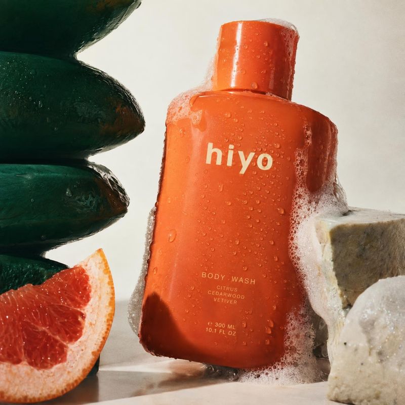 body wash personal care product photo: An orange body-wash bottle beaded with water and foam is styled with grapefruit, dark green soap and a stone-like prop a…