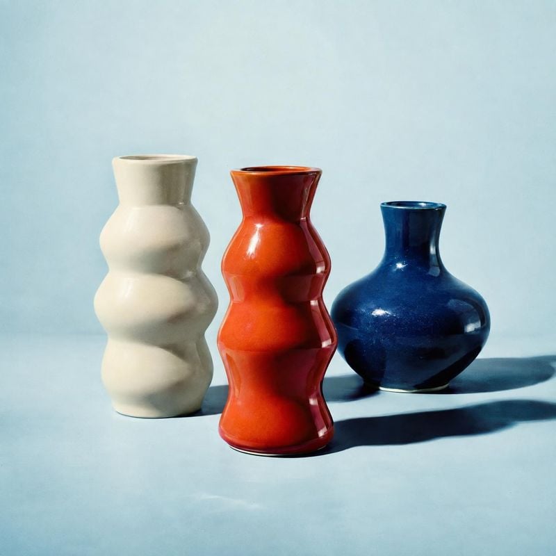 ceramic vases group lineup product photo: Three sculptural ceramic vases in cream, red, and deep blue stand against a pale blue studio backdrop, with crisp dir…