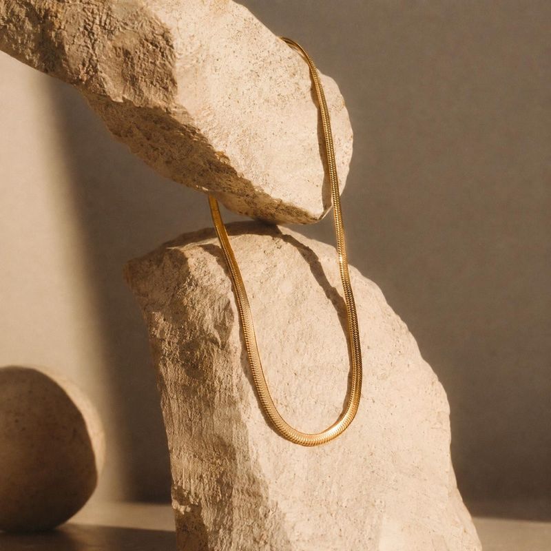 gold necklace jewelry product photo: A gold chain necklace is draped over pale sculptural stones, photographed in warm directional light for a quiet, refined m…