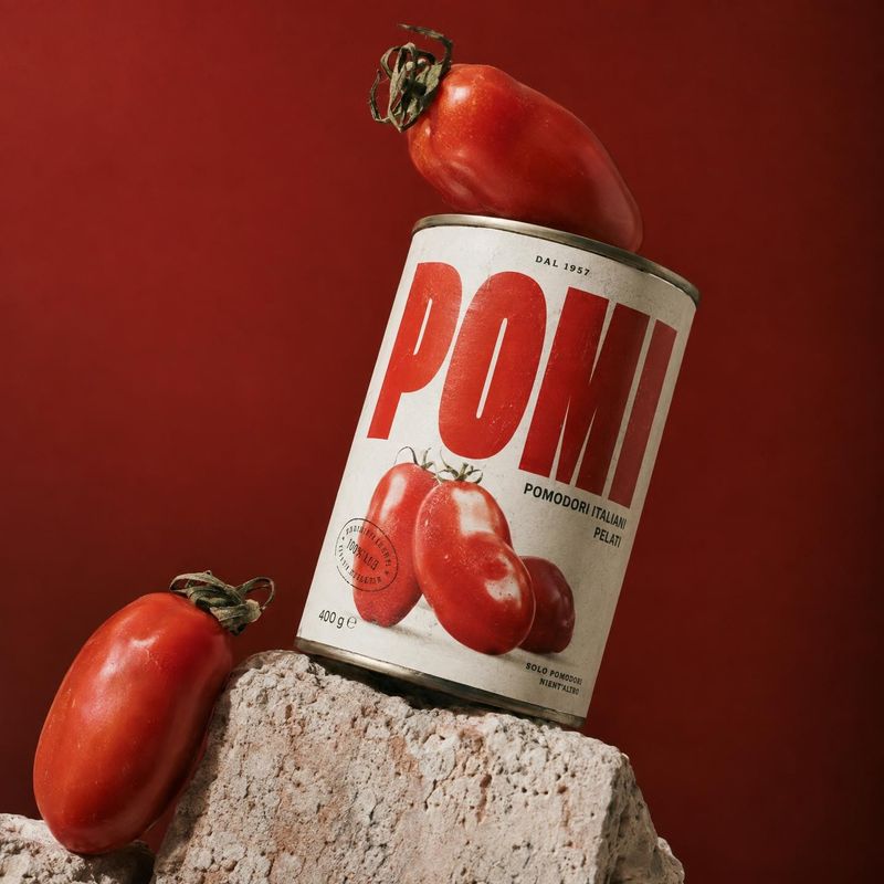 canned peeled tomatoes food and drink product photo: A can of peeled tomatoes leans against a rough stone block, framed by fresh tomatoes against a rich red st…