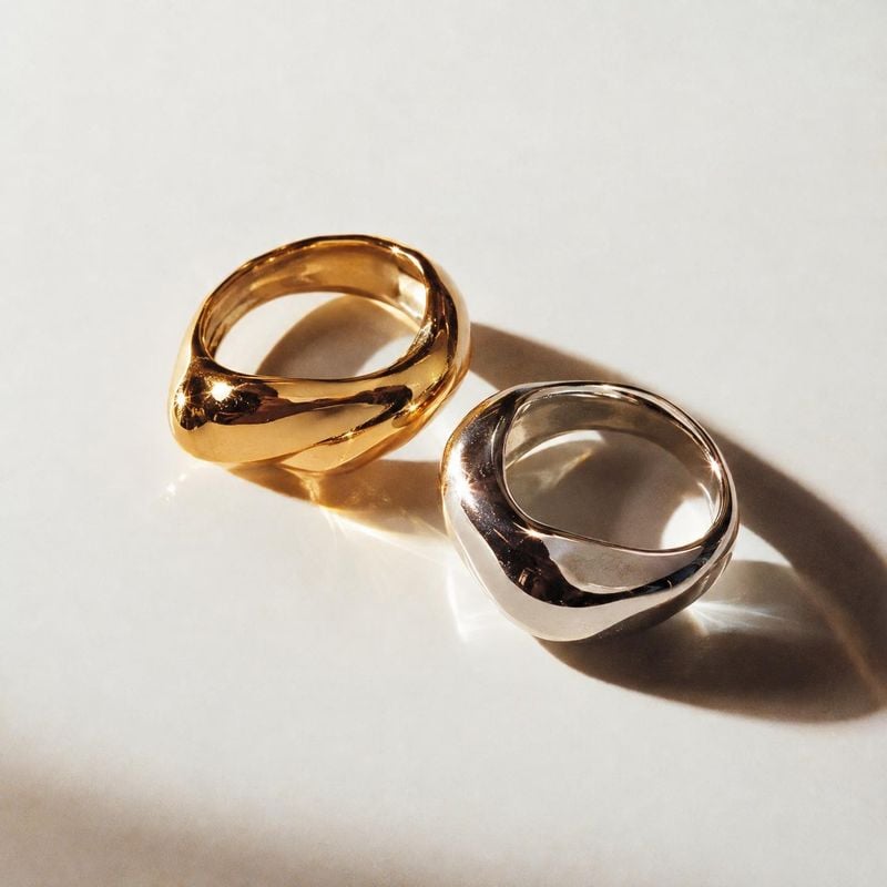 metal rings close-up detail product photo: Two polished gold-tone and silver-tone rings rest on a pale studio surface, their sculptural forms catching warm dir…