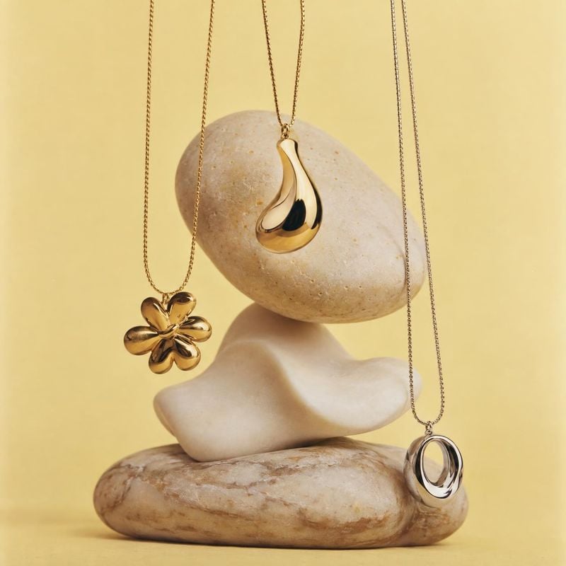 gold-tone necklaces jewelry product photo: Three sculptural gold-tone necklaces hang against a softly lit yellow backdrop, arranged with smooth natural stones…