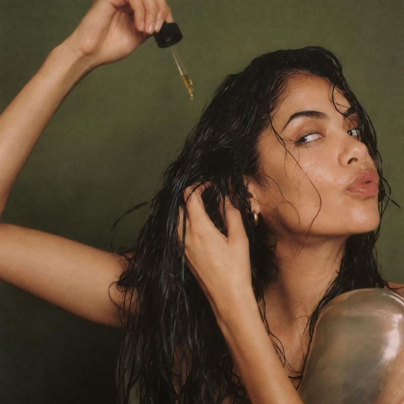 hair oil beauty close-up product photo: A woman with wet, dark hair applies oil from a dropper while touching her hair against a muted olive studio backdrop, c…