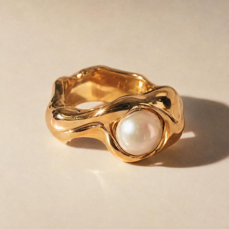 gold ring with pearl jewelry product photo: A sculptural gold ring set with a luminous pearl rests on a warm cream surface, lit by soft golden studio light for…