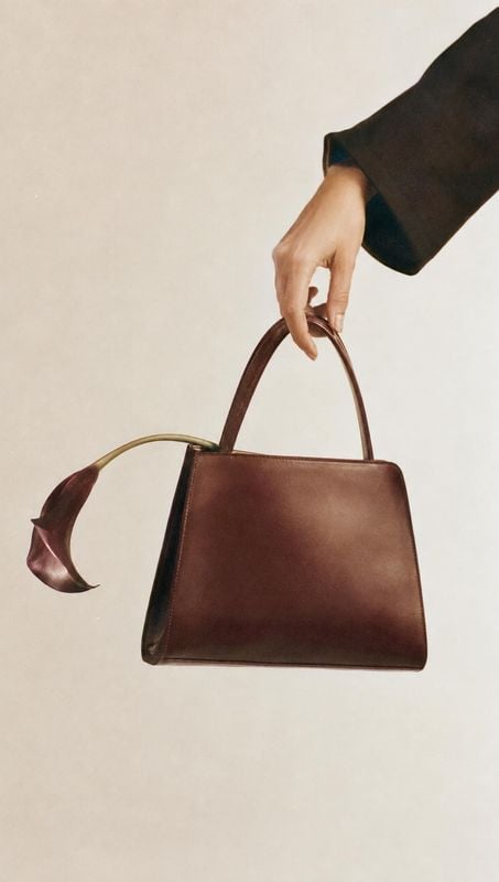 leather handbag product in hand product photo: A hand in a dark sleeve presents a structured burgundy leather handbag, with a sculptural calla lily beside it a…