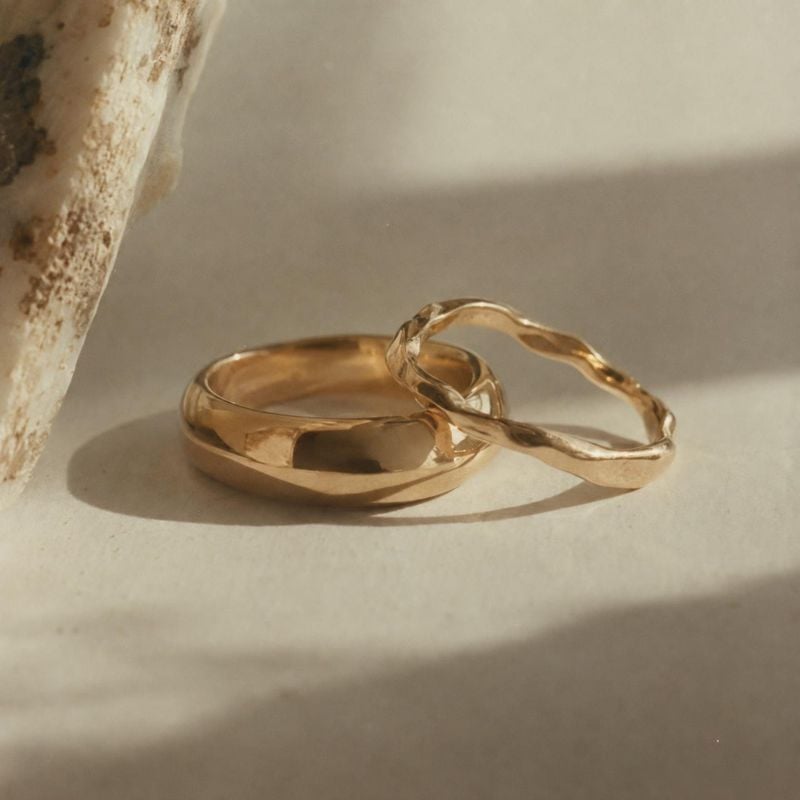 gold rings jewelry product photo: Two sculptural gold rings rest on a warm stone surface beside a weathered stone prop, lit by soft directional sunlight for a…