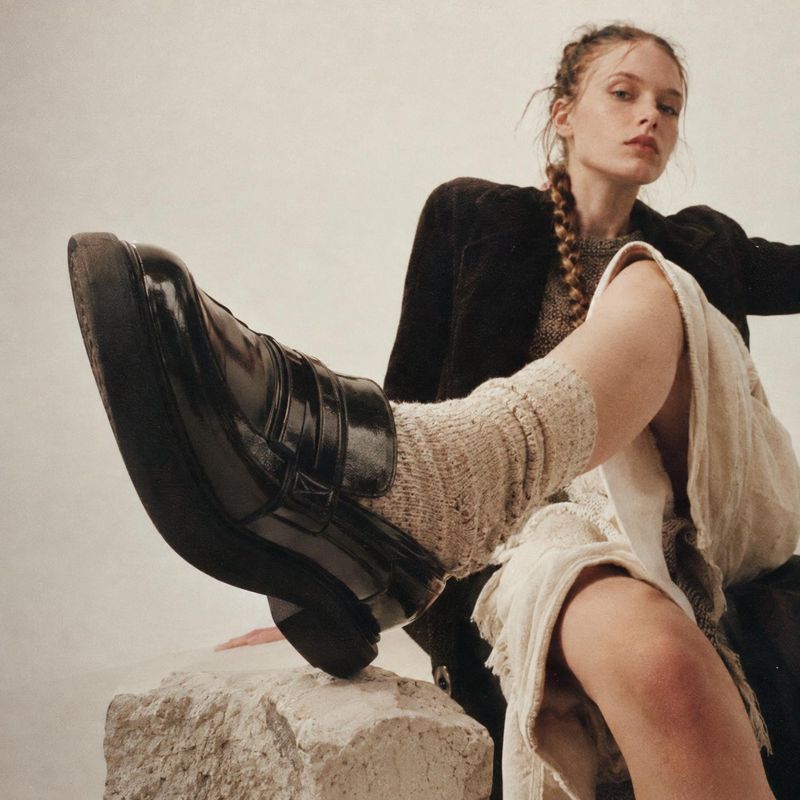 black leather boots model try-on product photo: A fashion model poses with one black boot extended toward the camera, styled with chunky knit socks and layered…